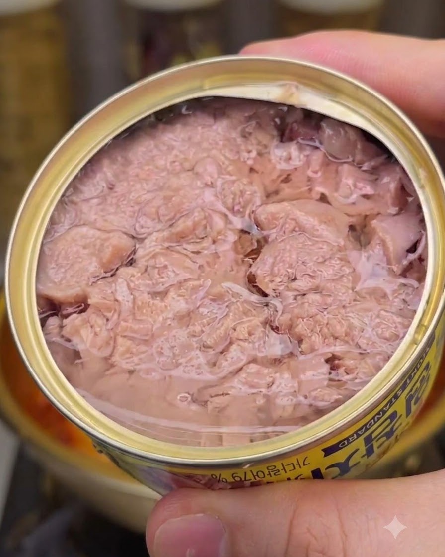 ピンク色のフレーク状のツナと缶汁が入った開いたツナ缶を、下に見える煮立った鍋の縁の近くで持ち上げているところ。