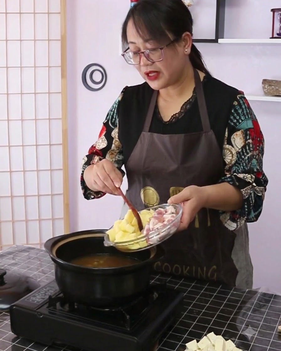 土豆和五花肉正从玻璃盘倒入盛有汤汁的黑色陶瓷砂锅中。