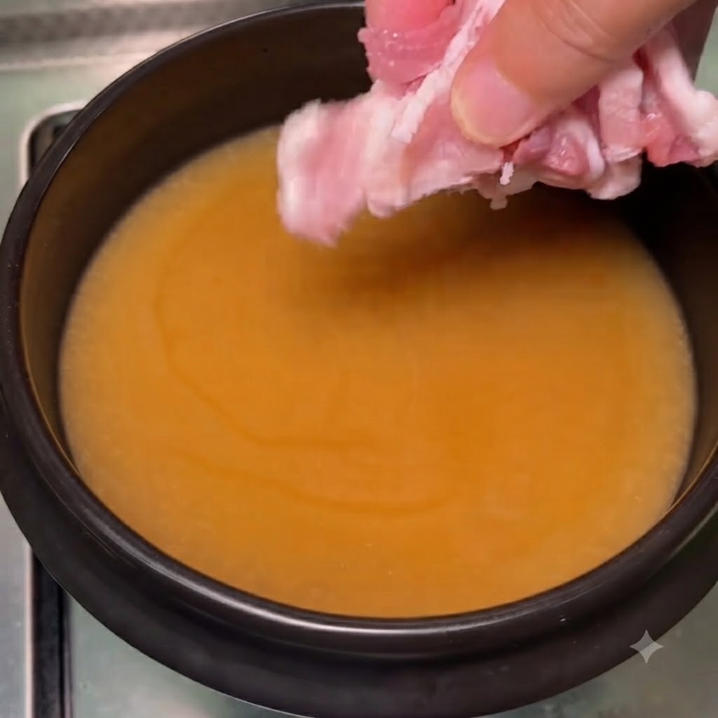 沸騰した味噌スープの中に、ピンク色の薄切り豚バラ肉を指で入れている様子。