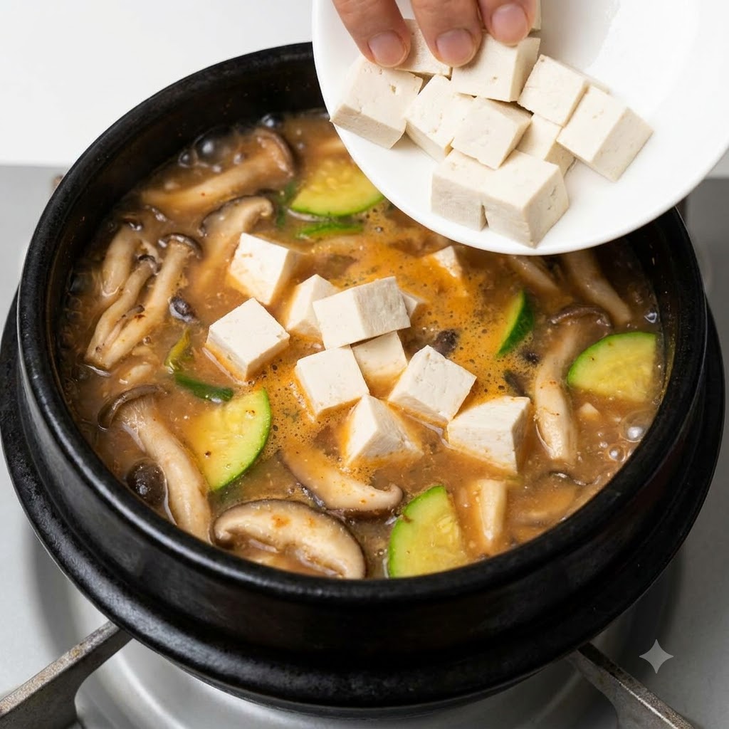 キノコやズッキーニが入った沸騰中の韓国味噌チゲに、小さな白い皿から角切りの白い豆腐が加えられている様子。