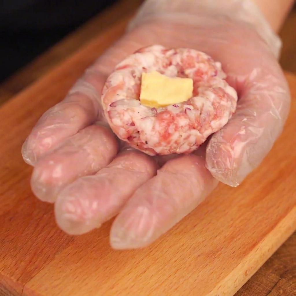 手袋をした手のひらで形を整えた豚ひき肉の凹みに、黄色いチーズのピースを置いているところ。