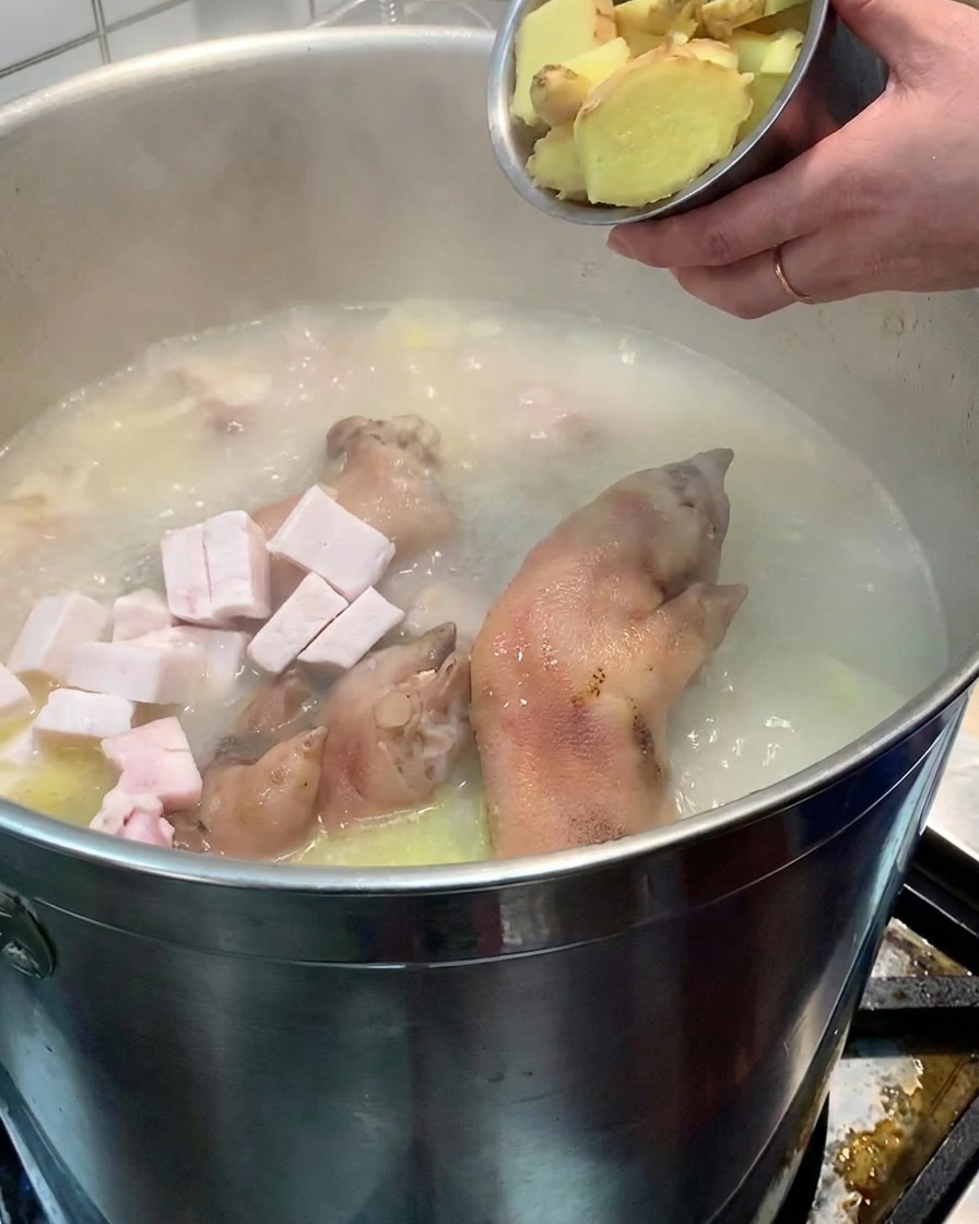 小さな金属製のボウルに入ったスライス生姜を、沸騰したスープと豚肉の入った大きな寸胴鍋に投入している人物。